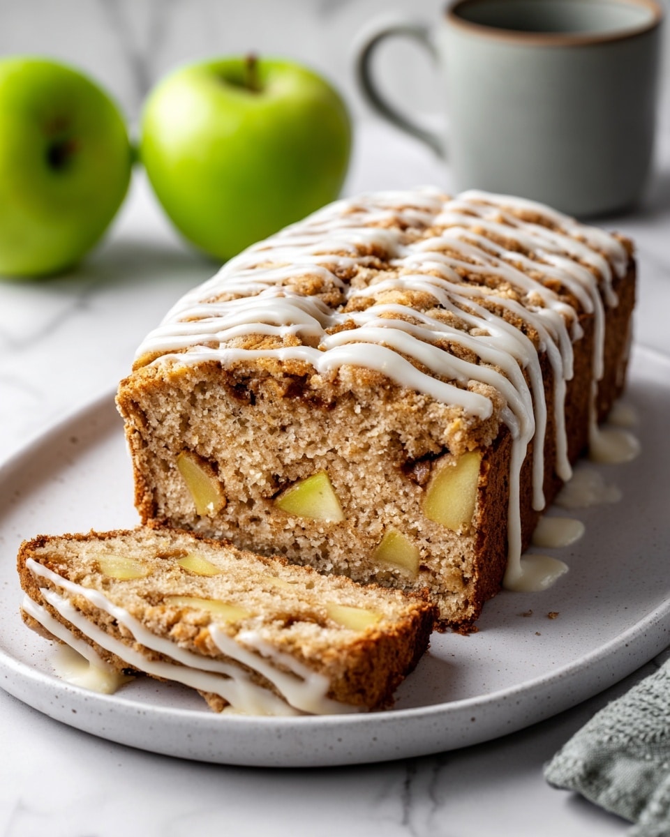 A loaf cake with two visible layers, cut partially to show its inside filled with small yellow apple pieces and cinnamon swirls, giving a mix of light beige, dark brown, and pale yellow colors. The top layer is covered with white icing drizzled in thin lines, some flowing slightly down the sides. The cake sits on a dark brown oval plate placed on a white marbled surface, and the background shows blurred green apples and a gray container. photo taken with an iphone --ar 4:5 --v 7