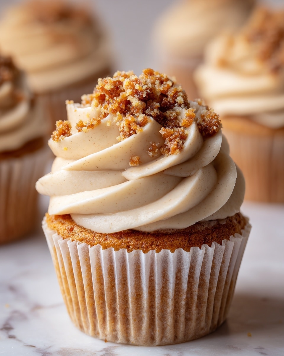 A close-up of a single cupcake with a light golden-brown base wrapped in a white paper liner, topped with a thick swirl of creamy beige frosting. On top of the frosting, there is a sprinkle of coarse brown crumbly streusel, adding texture and contrast. The background shows a soft-focus white marbled surface with more similar cupcakes blurred out behind the main one. The lighting highlights the smoothness of the frosting and the crumbly texture of the topping. photo taken with an iphone --ar 4:5 --v 7