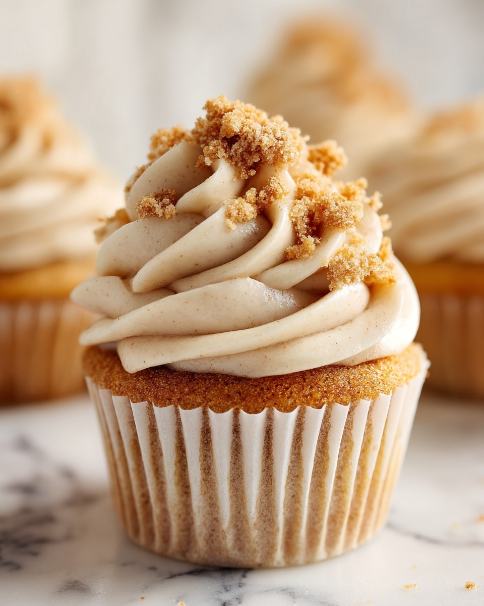A close-up of a single cupcake with a light brown base layer wrapped in a white cupcake liner. On top, there is a thick swirl of creamy, pale beige frosting with smooth texture, decorated with small clusters of crumbly, darker brown streusel bits scattered on the frosting. The cupcake sits on a white marbled surface, and a blurred background hints at more cupcakes of the same kind. The image is detailed, showing the soft texture of the cake and the creamy and crumbly topping. Photo taken with an iphone --ar 4:5 --v 7