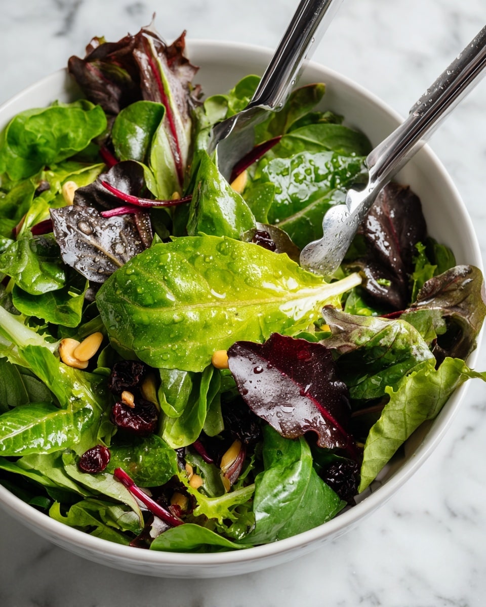 A close-up image shows a white bowl filled with a fresh green salad. The salad has layers of bright green lettuce leaves mixed with darker red and purple leaves, pine nuts sprinkled on top, and dried cranberries scattered throughout. A pair of silver tongs held by a woman's hand is lifting some green leaves from the bowl. The background is a white marbled texture. Photo taken with an iphone --ar 4:5 --v 7