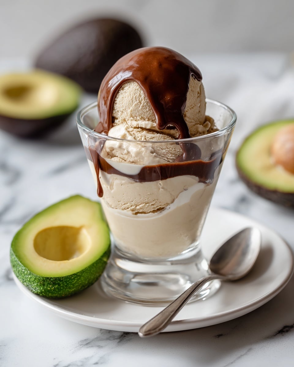 A clear glass cup sits on a white plate with a silver spoon beside it, all placed on a white marbled surface. Inside the cup are two main layers: the bottom layer is light brown ice cream filling the cup, and the top layer is a lighter brown scoop of ice cream. Over the scoop, thick dark chocolate sauce is drizzled in a smooth ribbon. Around the plate lie three avocado halves with green skin and pale yellow-green flesh, adding fresh color to the scene. Photo taken with an iphone --ar 4:5 --v 7