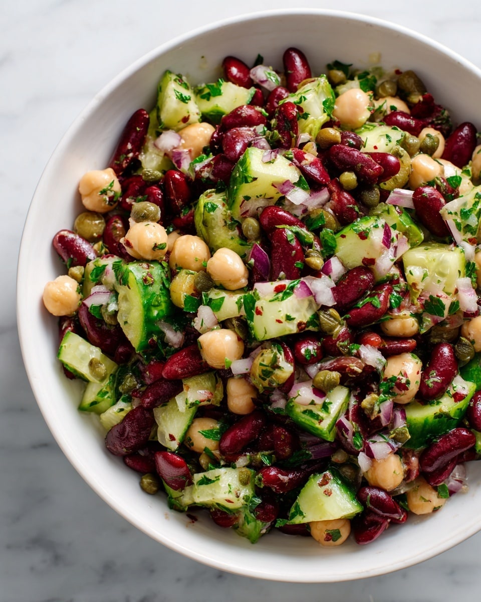 A bowl filled with a colorful mixed bean salad sits on a white marbled surface. The dish has three main layers: large dark red kidney beans, round light beige chickpeas, and bright green cucumber chunks with their skin on. These are scattered evenly throughout the bowl. Finely chopped purple-red onions and small green capers add texture and pops of color on top and within the mix. Fresh green herbs, likely parsley or cilantro, are sprinkled throughout, adding a hint of freshness visible in small leafy pieces. The bowl is white and simple with a slightly speckled rim, making the colors of the salad stand out vividly. photo taken with an iphone --ar 4:5 --v 7
