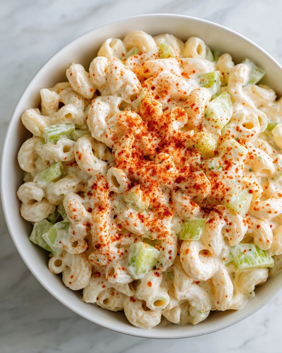 A white bowl filled with creamy macaroni salad, showing three main layers: the bottom layer of small curved macaroni noodles coated in a smooth, light beige mayonnaise dressing, the middle layer of bright green celery pieces mixed evenly throughout, and the top layer of the same creamy dressing sprinkled with a generous amount of reddish-brown paprika powder. The bowl sits on a white marbled surface, and the overall look is rich and fresh with a soft texture on the macaroni and crunchiness from the celery. photo taken with an iphone --ar 4:5 --v 7