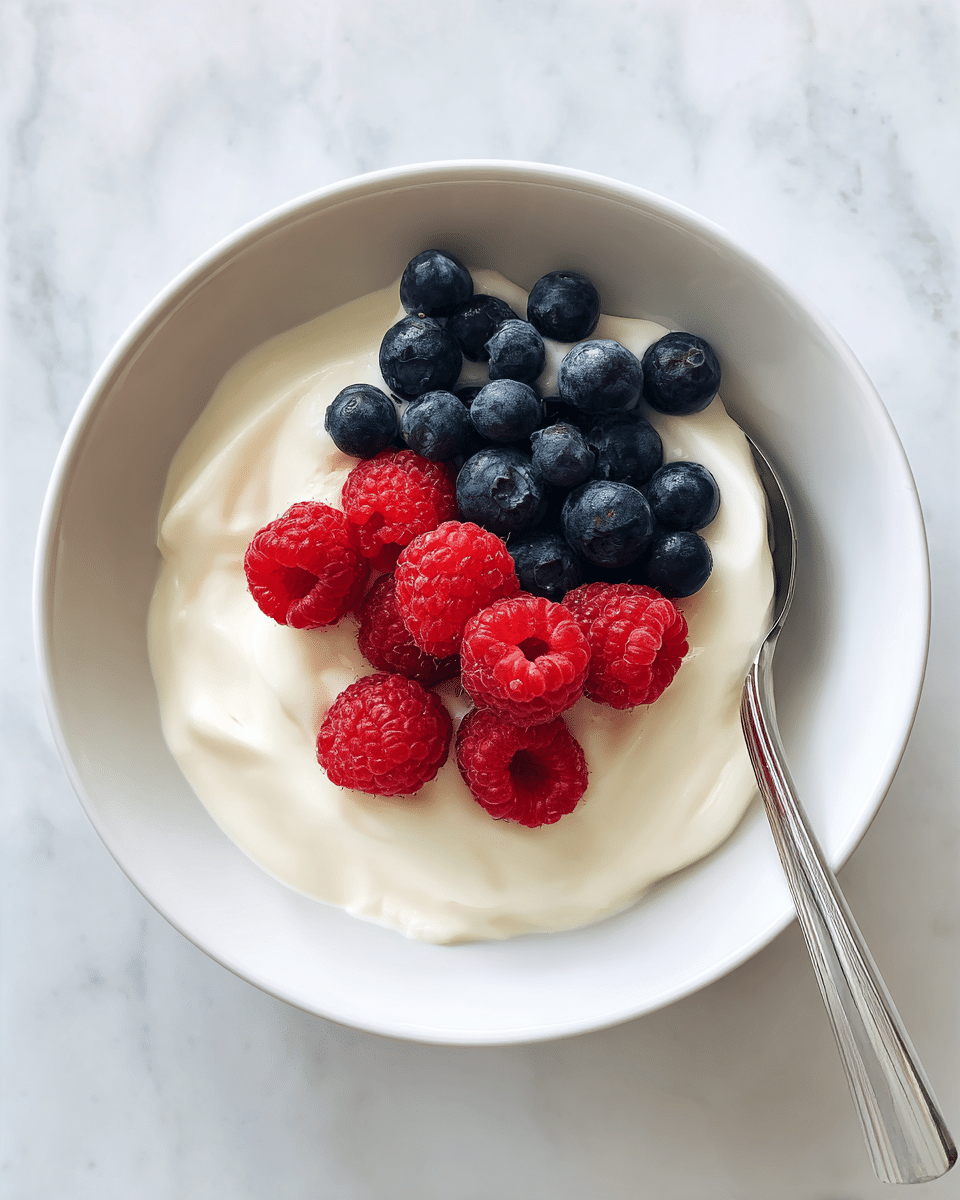 A white bowl filled with a thick, creamy white yogurt base, topped with a small pile of fresh red raspberries and dark blue blueberries placed at the center. A silver spoon is partly visible, standing in the bowl on the right side. The bowl sits on a white marbled surface. photo taken with an iphone --ar 4:5 --v 7