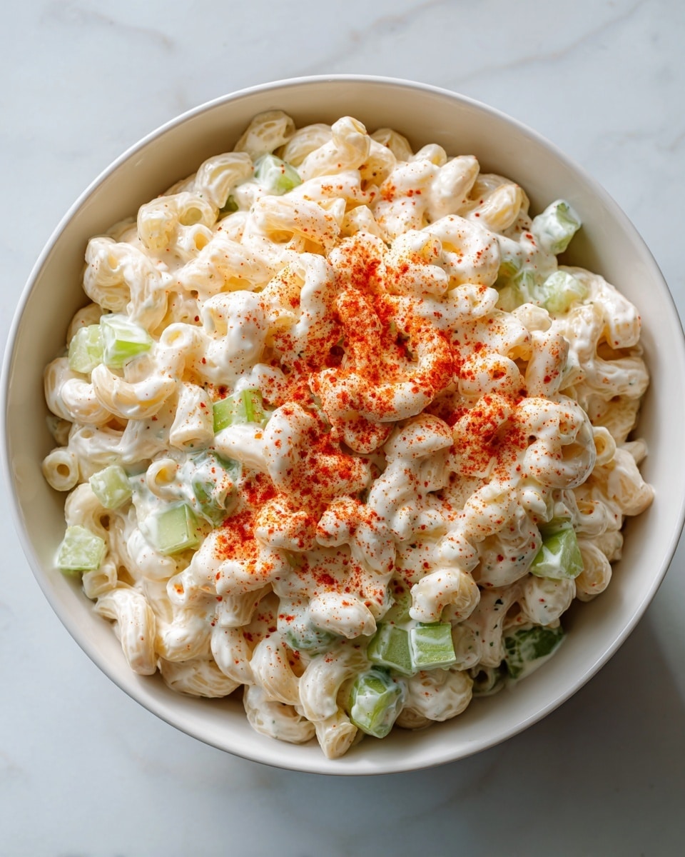 A white bowl filled with creamy macaroni salad layered with small elbow pasta coated in a shiny white sauce, mixed with light green chopped celery pieces evenly spread throughout. The salad is topped with a light sprinkle of red paprika powder concentrated in the center, creating a colorful contrast against the creamy white pasta. The bowl is set on a white marbled surface, giving a clean and fresh look to the image. photo taken with an iphone --ar 4:5 --v 7
