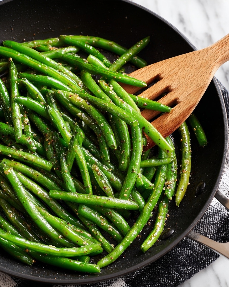 A close-up view of cooked green beans in a black cast iron pan shows a single layer of bright green beans with a slight shine, some beans showing light char marks, and small dark seasoning bits scattered throughout. A wooden spatula with a smooth texture and natural wood color is partially under the green beans, positioned diagonally from the bottom right corner toward the center of the pan. The pan rests on a white marbled surface with a cloth featuring a beige, black, and white checkered pattern partially visible in the top part of the image. Photo taken with an iphone --ar 4:5 --v 7