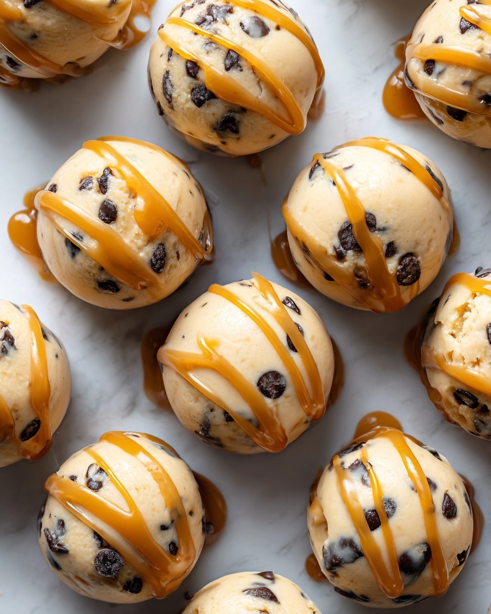 The image shows a close-up of small round frozen peanut butter yogurt bites arranged closely together. Each bite is a creamy light beige color with visible small dark chocolate chips mixed inside, and they are drizzled with smooth caramel sauce that adds a shiny golden brown layer on top. The bites have a slightly rough texture from the chocolate chips and a glossy layer from the caramel drizzle. The background is a white marbled surface, creating a clean and bright look. Photo taken with an iphone --ar 4:5 --v 7