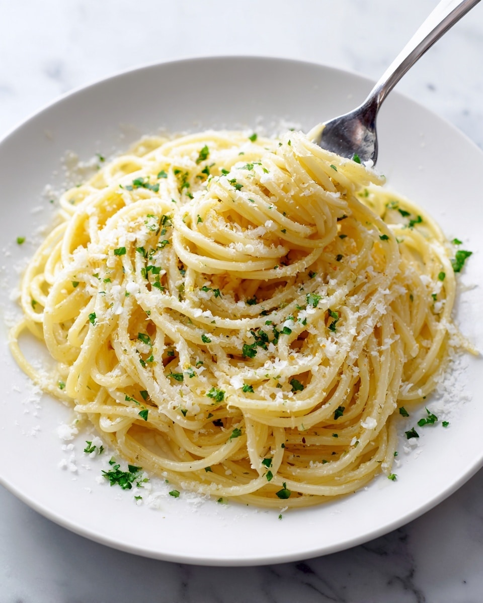 A close-up of a white plate filled with a single serving of spaghetti pasta, the noodles forming a loose mound with a pale yellow color and slightly glossy texture, sprinkled with fine white grated cheese and small pieces of bright green herbs scattered evenly on top. A silver fork, partially visible on the right side, is gathering some pasta strands. The plate sits on a white marbled surface, and the overall look is simple and fresh. photo taken with an iphone --ar 4:5 --v 7