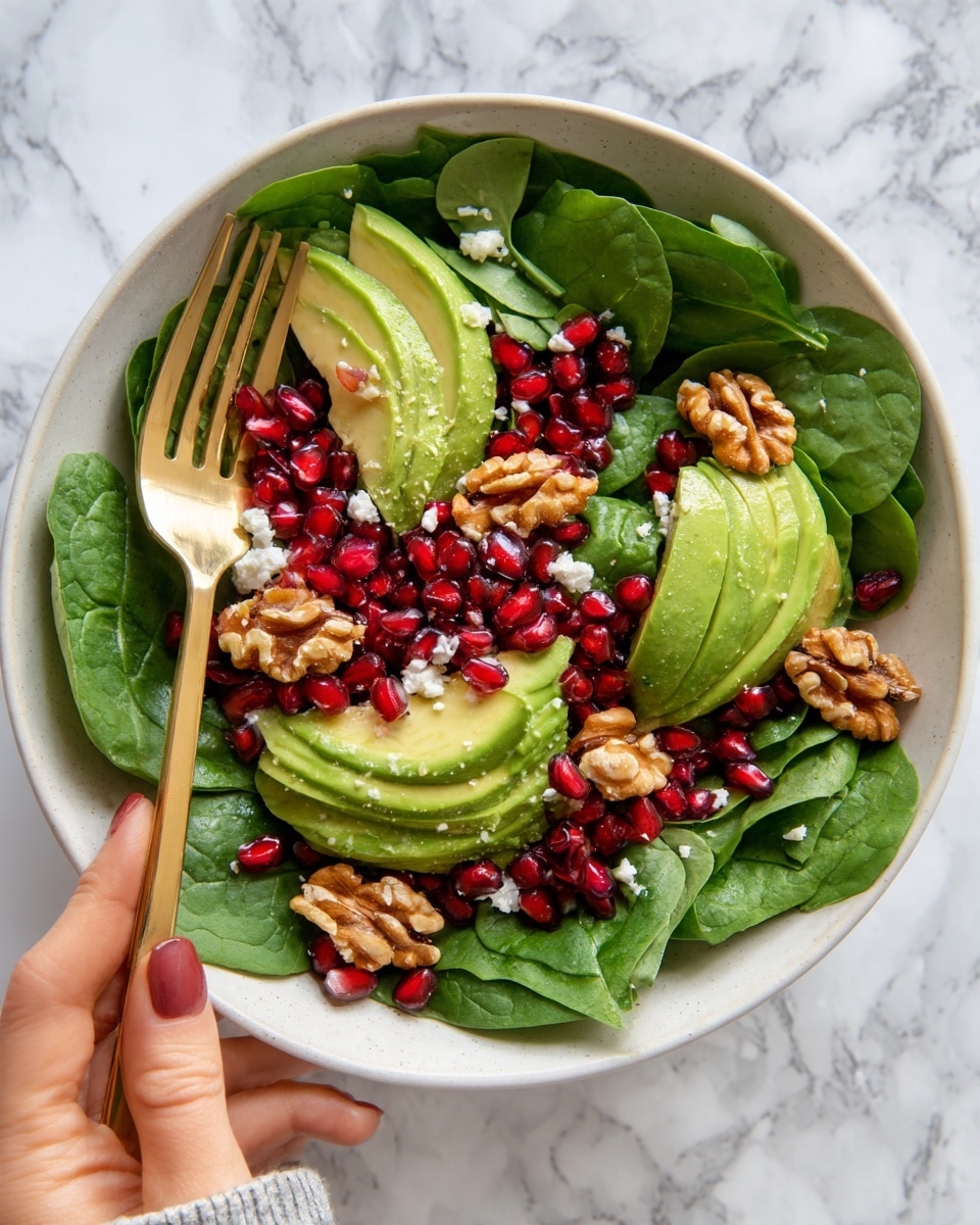 A fresh salad on a white plate with bright green spinach leaves forming the base layer. Sitting on top are several halves of ripe avocado with a smooth, light green inside and darker green edges. Scattered around the plate are small, crunchy brown walnut pieces and bright red pomegranate seeds that add color contrast. White crumbled cheese is sprinkled over everything, giving a soft texture. The plate rests on a white marbled surface with a red cloth partially visible under one side. Photo taken with an iphone --ar 4:5 --v 7