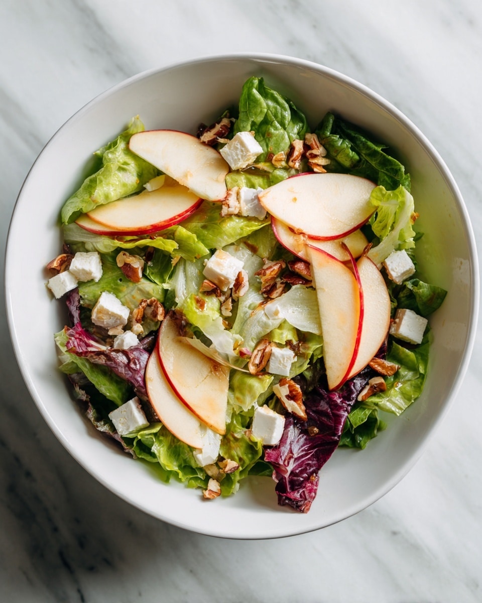The image shows a bowl of fresh salad with three clear layers. The bottom layer is green lettuce leaves that look crisp. Above the lettuce, there are thin slices of red-skinned apples arranged evenly across the bowl. The top layer has small white chunks of cheese and scattered pieces of light brown walnuts, giving a crunchy texture. The bowl is white, and the background is a white marbled surface. Photo taken with an iphone --ar 4:5 --v 7