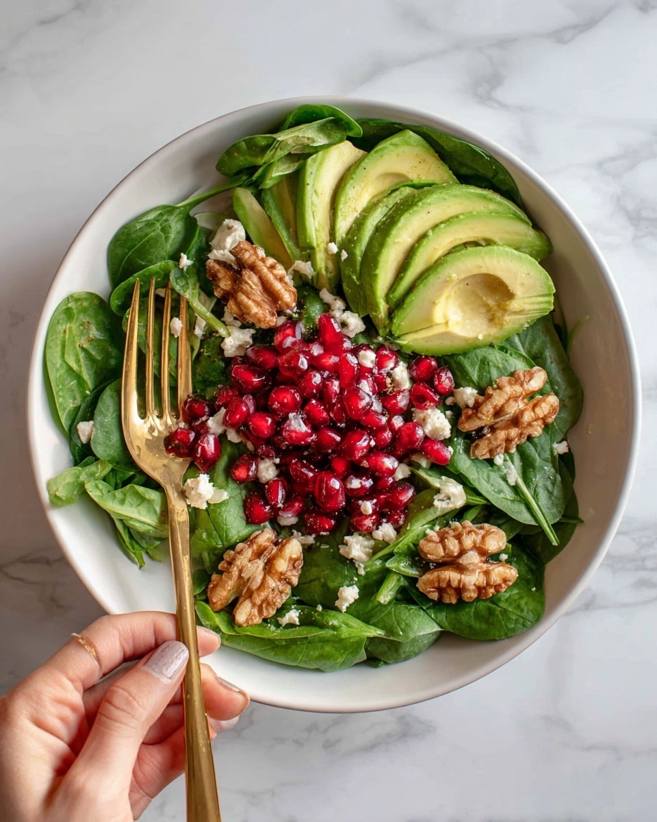 The image shows a fresh salad served in a white bowl filled with dark green spinach leaves as the base layer. On top, there are bright red pomegranate seeds scattered evenly, adding a jewel-like touch. Halved avocado pieces with smooth light green flesh and darker green skin are placed around the bowl. Crunchy walnut halves, light brown in color with textured surfaces, are distributed evenly. Small white crumbles of cheese are sprinkled throughout. A woman's hand holding a golden fork is seen on the edge of the bowl, ready to take a bite. The bowl rests on a white marbled surface. Photo taken with an iphone --ar 4:5 --v 7